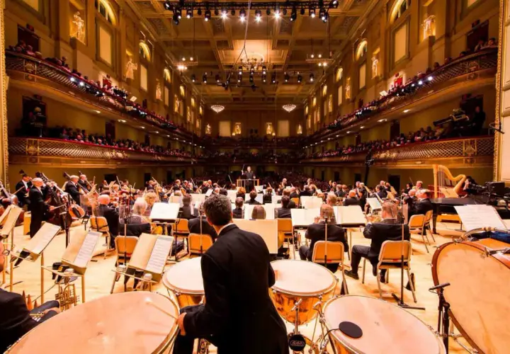 Un orchestre symphonique interprète de la musique classique moderne dans une salle de concert ornée, avec des musiciens jouant de divers instruments.
