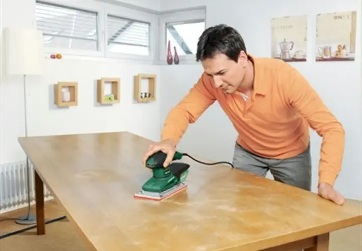Un homme utilise une ponceuse vibrante ou excentrique pour rénover une table en bois.