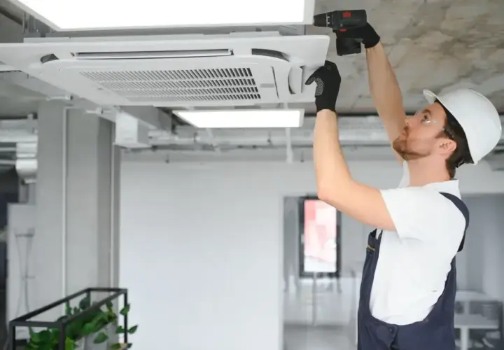 Instalator w kasku montuje klimatyzator kasetonowy na suficie.