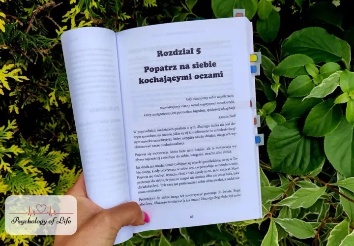 Otwarta książka "Popatrz na siebie kochającymi oczami" w otoczeniu zieleni. Tekst mówi o walce z negatywną samokrytyką i brakiem akceptacji siebie.