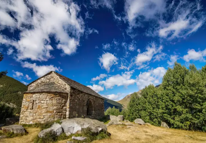 Pequeña ermita de piedra en las rutas del Vall de Boí, bajo un cielo azul con nubes algodonosas.