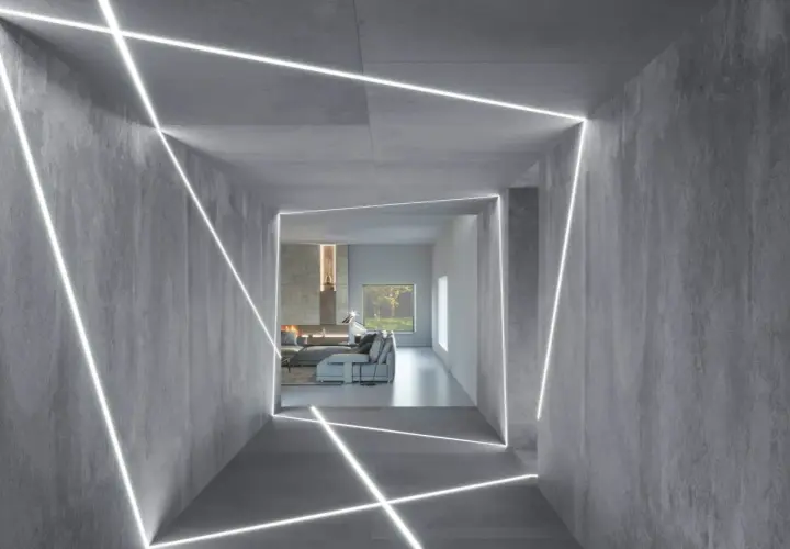Nowoczesny korytarz z betonowymi ścianami i geometrycznym podświetleniem ściany od góry. W tle salon z kominkiem i kanapą.