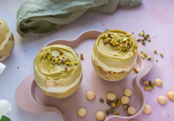 Dwa pucharki z deserem: krem pistacjowy z mascarpone, przełożony biszkoptami i bitą śmietaną, posypany pistacjami.