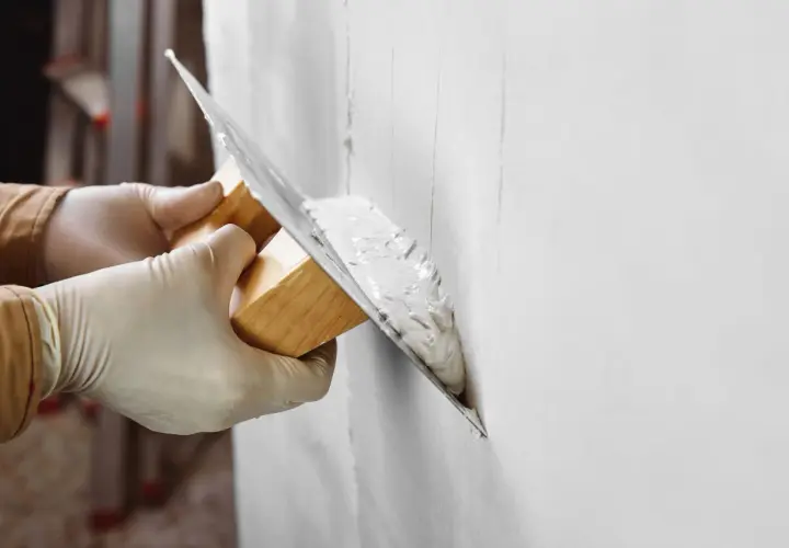 Manos con guantes aplican yeso a una pared. Pregúntate: cuanto tarda el yeso en secar para poder pintar.