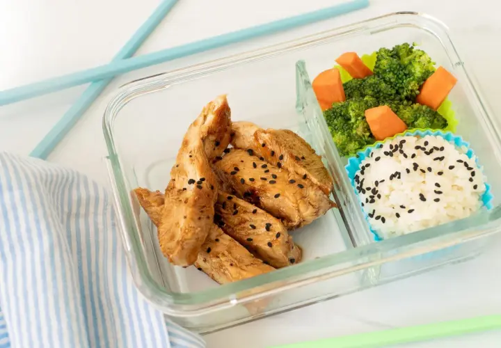 A delicious chicken bento box with teriyaki chicken strips, white rice sprinkled with black sesame seeds, and steamed broccoli with carrots.