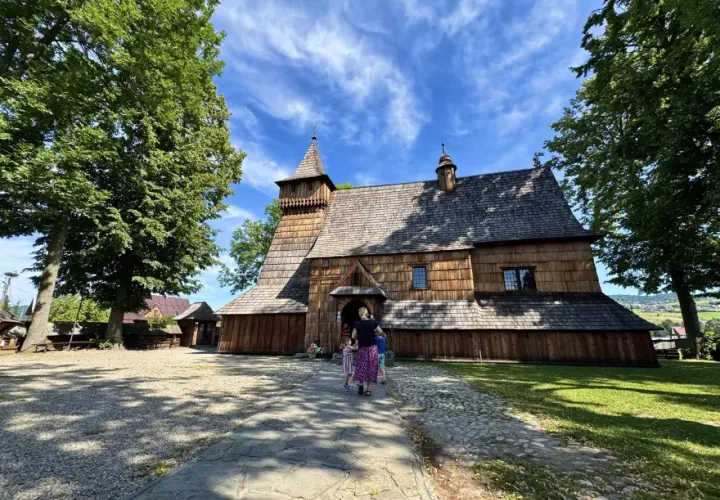 Drewniany kościół w Dębnie, z charakterystyczną wieżą i gontowym dachem, otoczony zielenią.