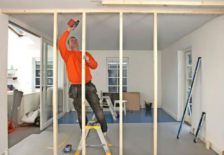 Man i orange tröja monterar träreglar för en ny vägg med skruvdragare.