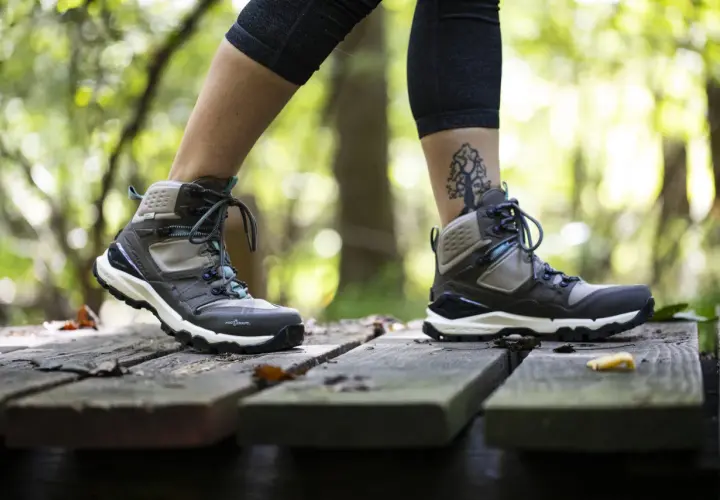 A person walks on a wooden path in the forest, wearing hiking boots. One foot half size bigger than the other, with a tree tattoo on the ankle.