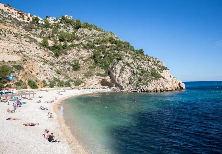 Gente disfrutando del sol en una playa de guijarros en la **Costa Blanca**, con aguas turquesas y un acantilado rocoso.