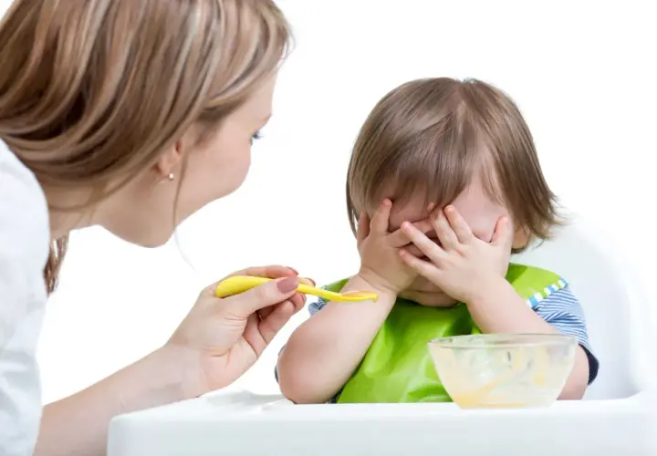 Mama próbuje nakarmić malucha, ale niemowlę nie chce jeść, zakrywając buzię rączkami.