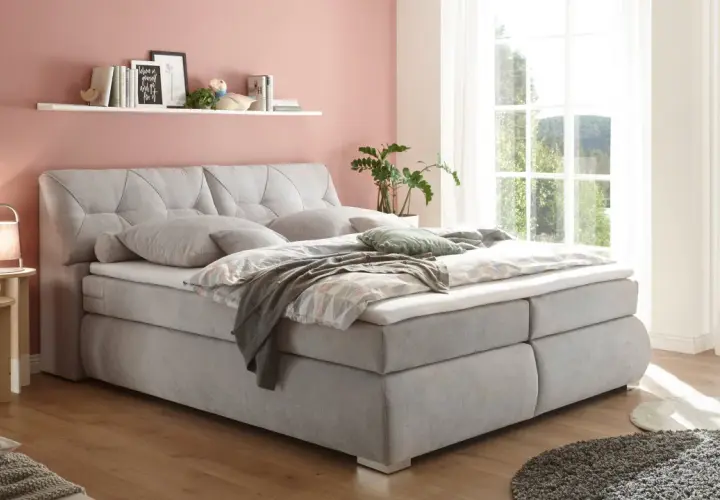 Ein hellgraues Boxspringbett mit Kissen und Decken lädt zum Entspannen ein. Die Wand in Rosé und das Fenster mit Blick ins Grüne schaffen eine harmonische Atmosphäre für ein **Schlafzimmer Feng Shui**.