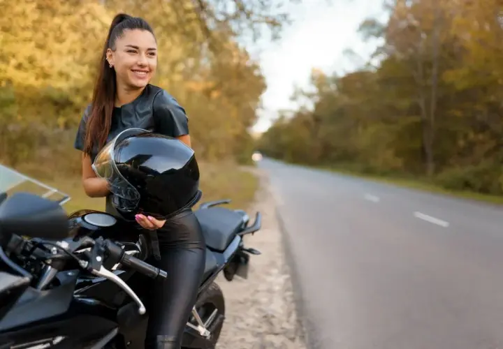 Uśmiechnięta kobieta w skórzanym stroju trzyma kask obok czarnego motocykla. Idealne motory dla kobiet na jesienną przejażdżkę.