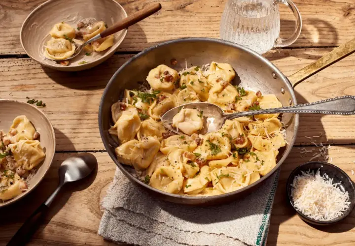 Herzhafte Tortellini Carbonara, garniert mit Petersilie und Parmesan, serviert in einer Pfanne und Schalen auf Holztisch.