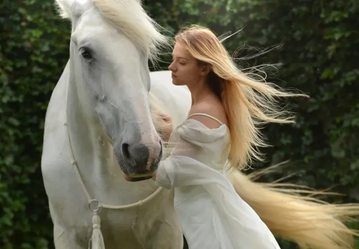 Una joven rubia con un vestido blanco se inclina hacia un majestuoso caballo blanco. Es como soñar con caballos, una escena etérea.
