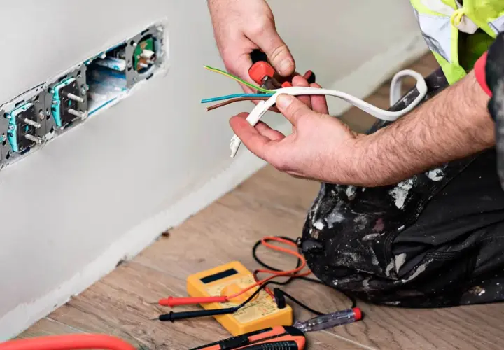 Elektryk przygotowuje kabel czarny, brązowy, niebieski do podłączenia gniazdek. Narzędzia i miernik leżą na podłodze.