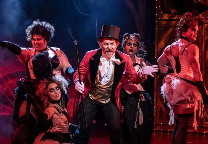 Scenen från Moulin Rouge! visar en cirkusdirektör med en cylinderhatt och en gyllene väst, omgiven av artister i glittrande kostymer. Handlar om kärlek och frihet i Paris.