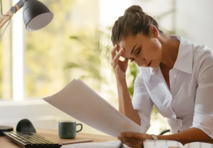 Frau mit Kopfschmerzen blickt auf Dokumente, die sie vielleicht für eine verbindliche Auskunft beim Finanzamt benötigt.