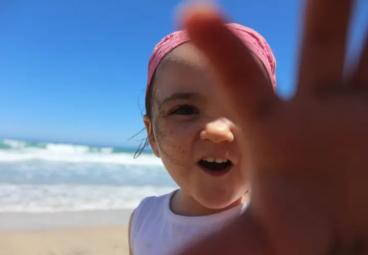 Niña con bandana rosa y arena en la cara, disfrutando de sus viajes con niños en la playa.
