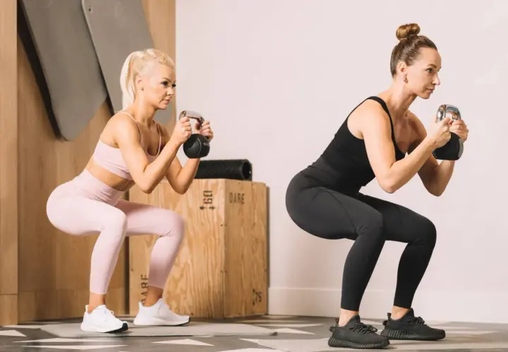 Dwie kobiety wykonujące przysiady z kettlebellami w siłowni.