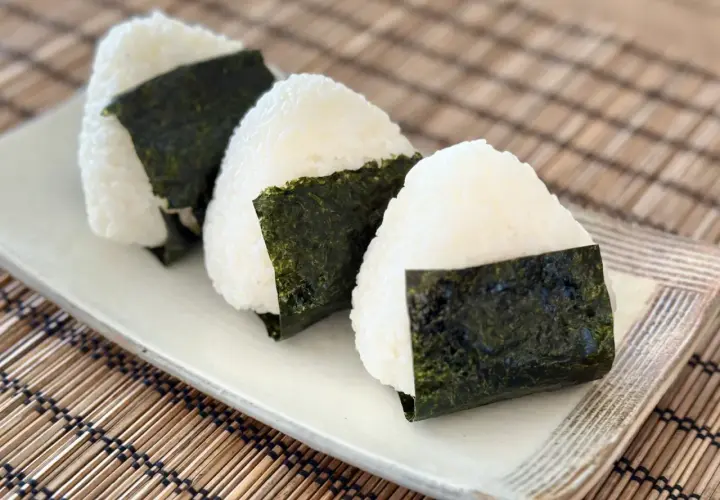 Three triangular onigiri, made with fluffy sushi rice and wrapped in dark green nori seaweed, sit on a white plate.