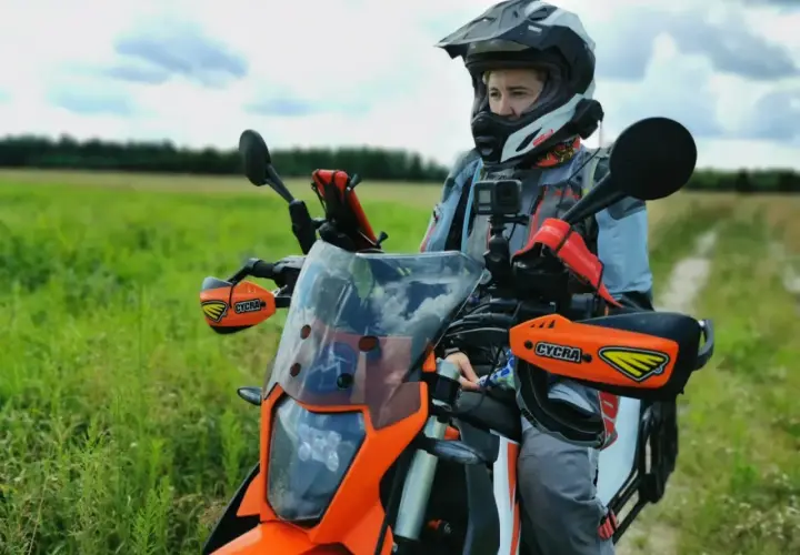 Motocyklista w kasku siedzi na pomarańczowym motocyklu w terenie, na tle zielonego pola i nieba.