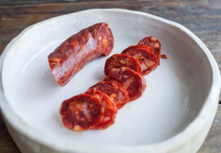 Ein Stück und Scheiben einer würzigen Salsiccia Wurst liegen in einer weißen Schale auf einem Holztisch.