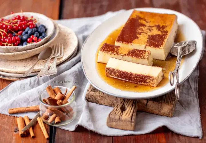 Pyszny creme caramel na talerzu, polany syropem. Obok miseczka z owocami i laska cynamonu.