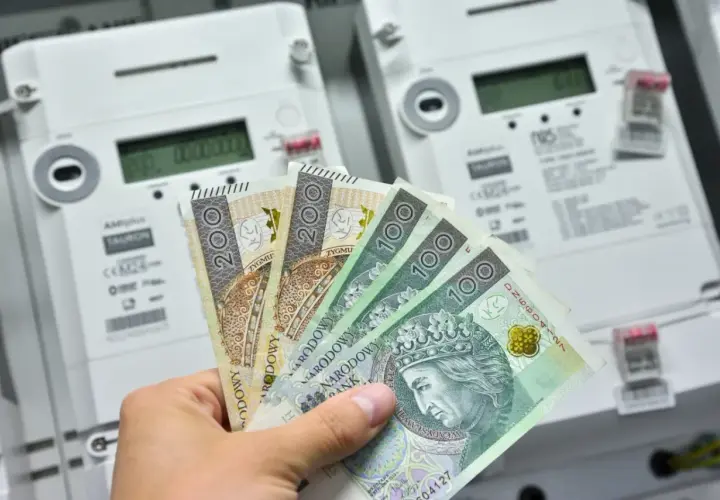Dłoń trzyma polskie banknoty przed licznikami energii elektrycznej. Zastanawiasz się, ile prądu zużywa zamrażarka miesięcznie?
