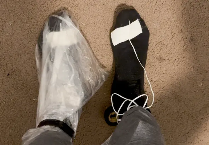 A person's feet are shown, one covered in a plastic bag, the other in a black sock with a white string and tape. These are makeshift vapor barrier liner socks for a DIY project.