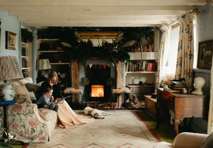 Cozy cottagecore house scene with a woman and child reading by a fireplace, dogs resting, and festive Christmas decorations.
