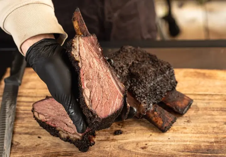 Une main gantée présente une côte de bœuf fumée, tendre et juteuse, sur une planche en bois.