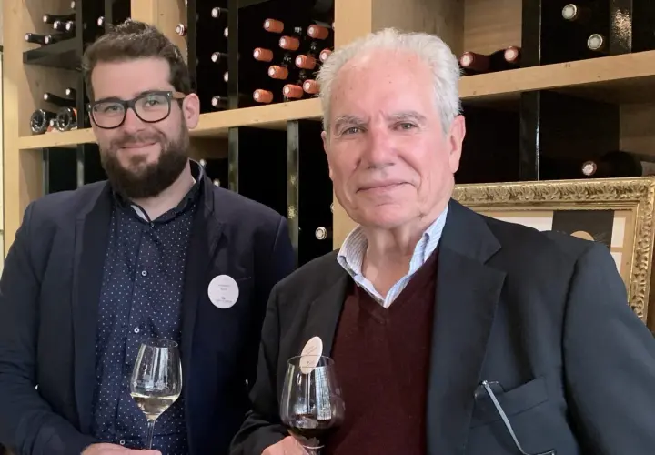 Deux hommes dégustent du vin devant des étagères remplies de bouteilles. L'un porte un badge "Domaine Rozel - Grignan-les-Adhémar avis".