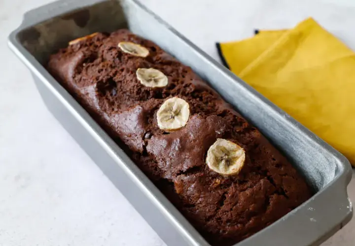 Plumcake banana e cioccolato appena sfornato, decorato con fettine di banana essiccata, in uno stampo metallico.