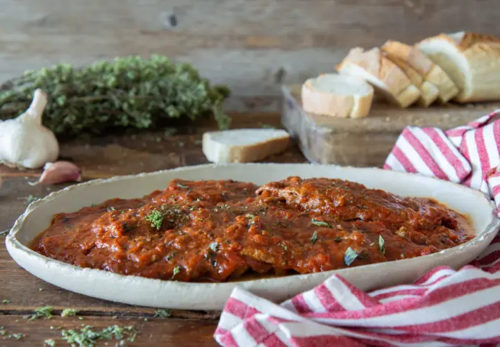 Herzhaftes Carne alla Pizzaiola in Tomatensauce, garniert mit Kräutern, neben Knoblauch, Brot und einem rot-weiß gestreiften Tuch.