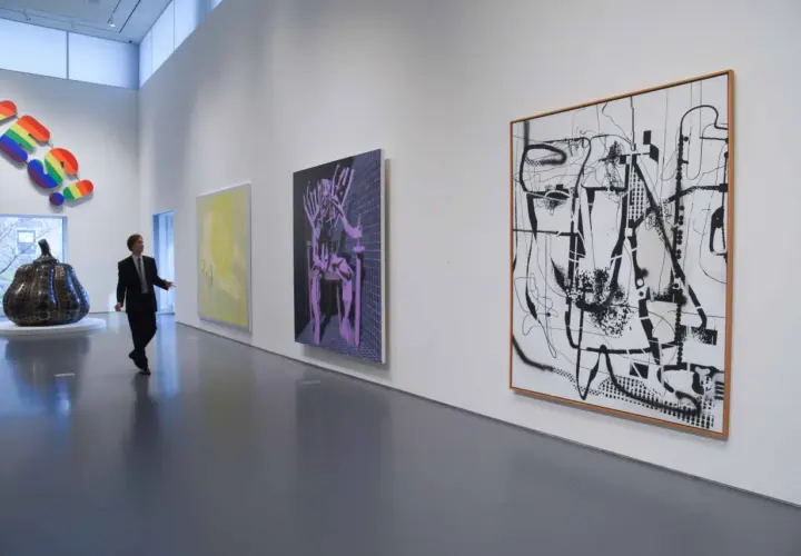 Un homme en costume observe une **analyse d'une œuvre d'art contemporain** dans une galerie. Des peintures abstraites et une sculpture colorée ornent les murs blancs.
