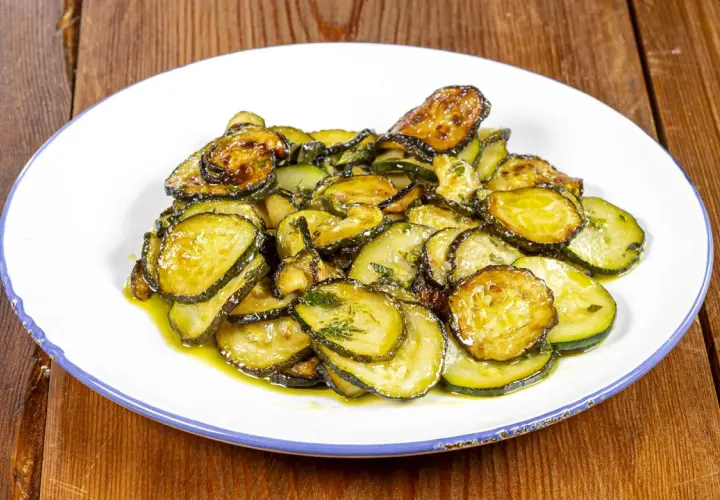 Piatto di zucchine alla poverella, fette sottili e dorate condite con olio e erbe aromatiche, su un piatto bianco con bordo blu.