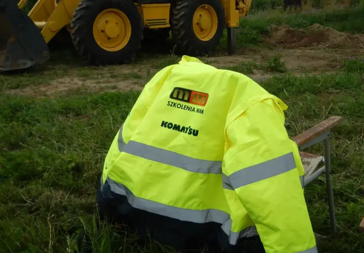 Kamizelka z logo Komatsu i napisem "Szkolenia MM" leży na krześle obok koparki. Idealna na testy na koparki jednonaczyniowe.