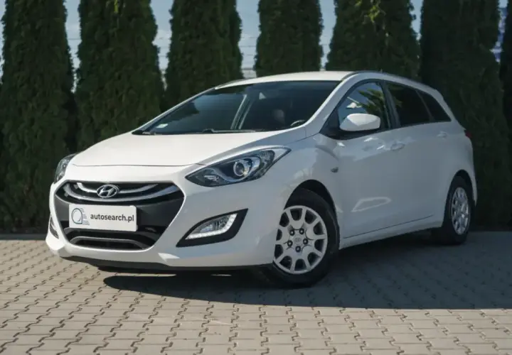 Biały Hyundai i30 1.4 benzyna na tle zielonych drzew, zaparkowany na kostce brukowej.
