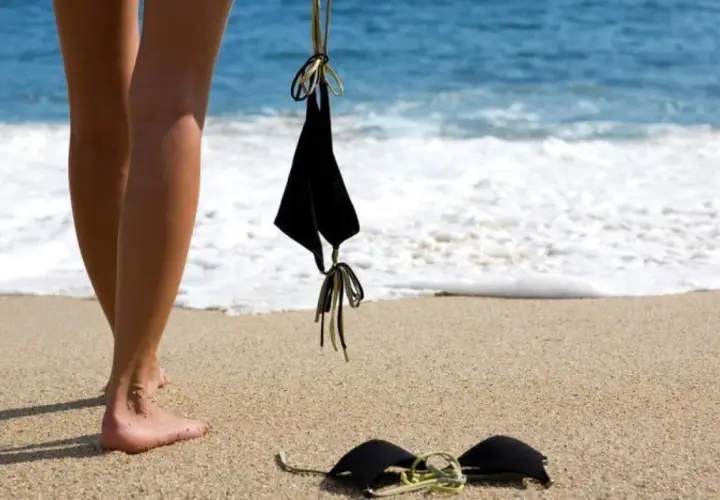 Nogi na plaży, obok leżą czarne bikini. Słońce grzeje, a fale obmywają brzeg. Czas na kąpiel dla golasy.