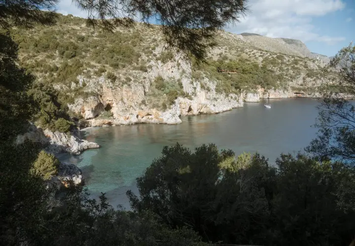 A serene view of Baia degli Infreschi, a secluded cove with clear turquoise waters, rocky cliffs, and lush greenery. A lone sailboat drifts peacefully in the bay.