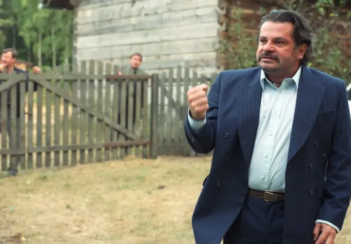 Mężczyzna w garniturze, jakby z filmu "To ja, złodziej", pokazuje pięść. Za nim przez płot wyglądają inni.