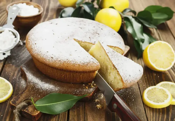 Una fetta di torta al limone morbidissima e profumatissima benedetta, cosparsa di zucchero a velo, con limoni freschi e foglie verdi su un tagliere di legno.