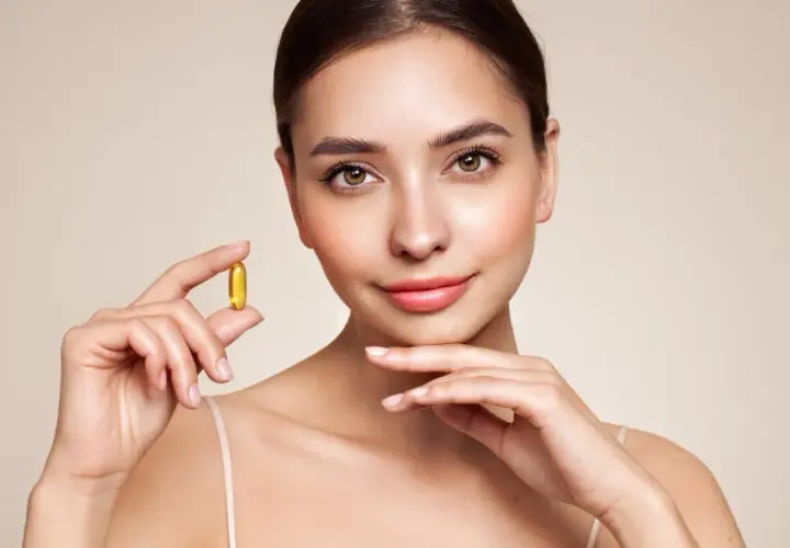 Mujer sonriente sostiene una cápsula dorada de vitamina e, símbolo de cuidado y bienestar para una piel radiante.