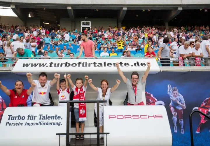 Zwei Kinder und zwei Erwachsene jubeln auf einer Tribüne vor einem Banner mit Bullen und einem Läufer.