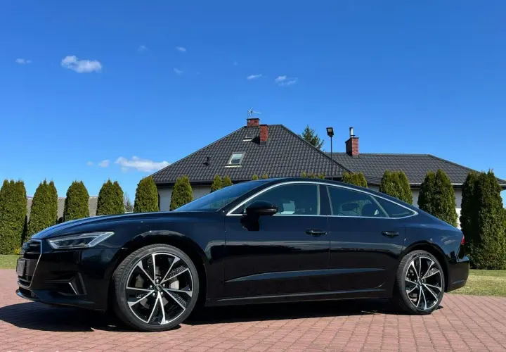 Elegancki czarny Audi A7 C8 lśni w słońcu na tle domu i zielonych drzew.