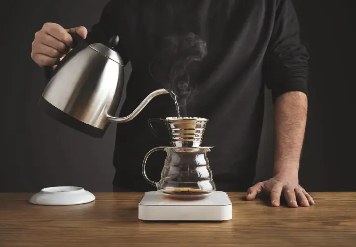 Mężczyzna przelewa gorącą wodę z czajniczka do drippera Kalita, przygotowując aromatyczną kawę.