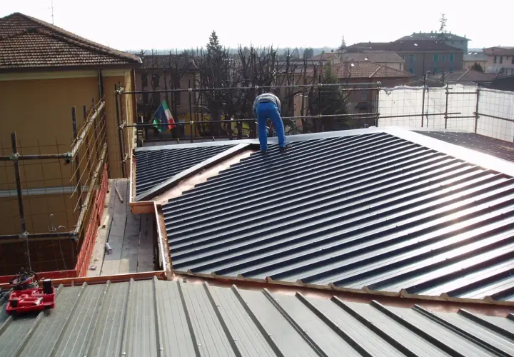 Operaio su tetto con muricci e tavelloni in fase di posa. Scaffolding e bandiera italiana sullo sfondo.