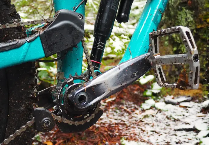 Close-up of a muddy mountain bike drivetrain with a teal frame, showing the crankset, chain, and pedal.