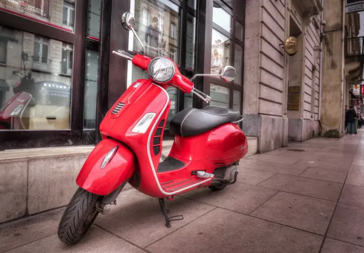 Czerwony skuter Vespa, idealny do zdobycia **prawo jazdy AM**, stoi na chodniku przed budynkiem.