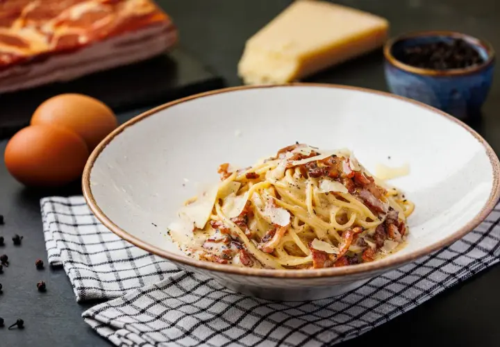 Un plato de spaghetti carbonara cremoso, con trozos de panceta crujiente y queso rallado. Ingredientes frescos como huevos, queso y pimienta negra completan la escena.